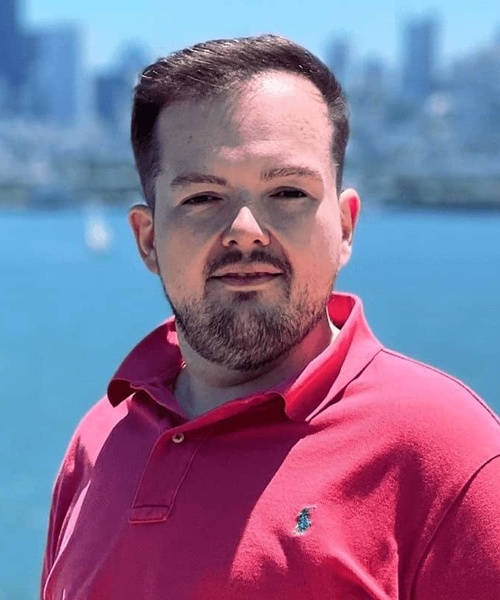 A man in a red polo shirt standing before a body of water with a city skyline in the background.