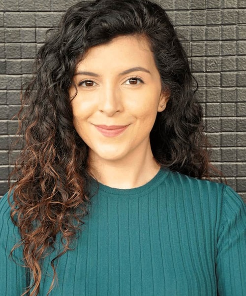 A woman with curly hair smiling at the camera against a brick wall background.