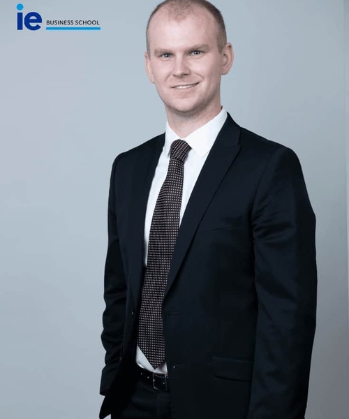 A professional portrait of a man in a business suit with a confident smile, standing against a grey background.