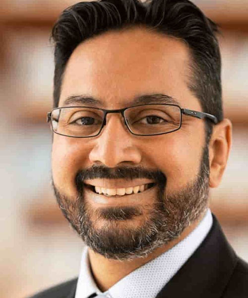 Portrait of a smiling man with a beard wearing glasses and a suit.