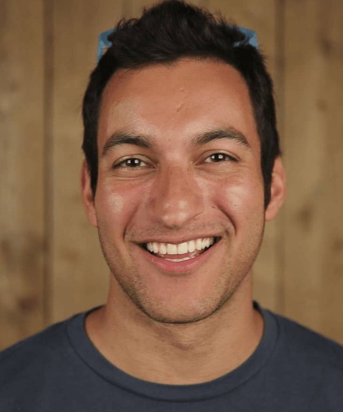 A portrait of a smiling man with dark hair wearing a blue t-shirt, set against a wooden background.