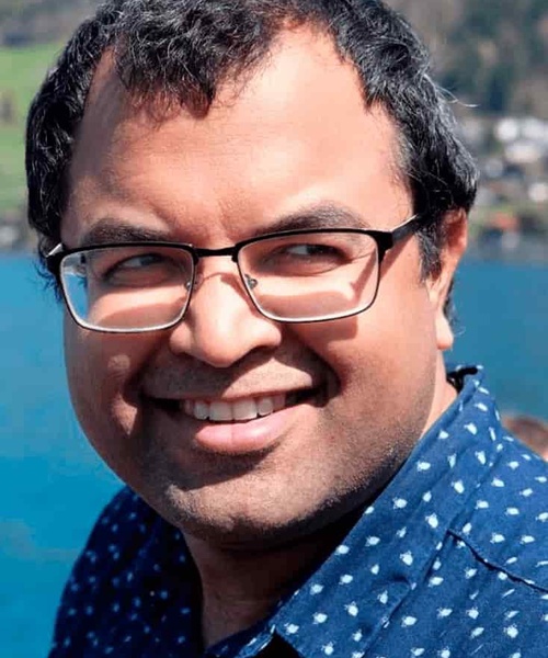 A smiling man with glasses wearing a blue polka dot shirt, with a blue water body and hills in the background.