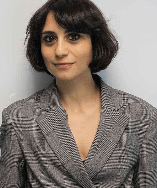 A woman with short dark hair wearing a gray blazer stands against a plain background.