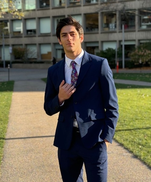 A young man in a blue suit adjusting his tie, standing outdoors with buildings in the background.