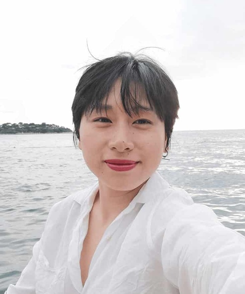 A young woman in a white blouse taking a selfie with the ocean and overcast sky in the background.