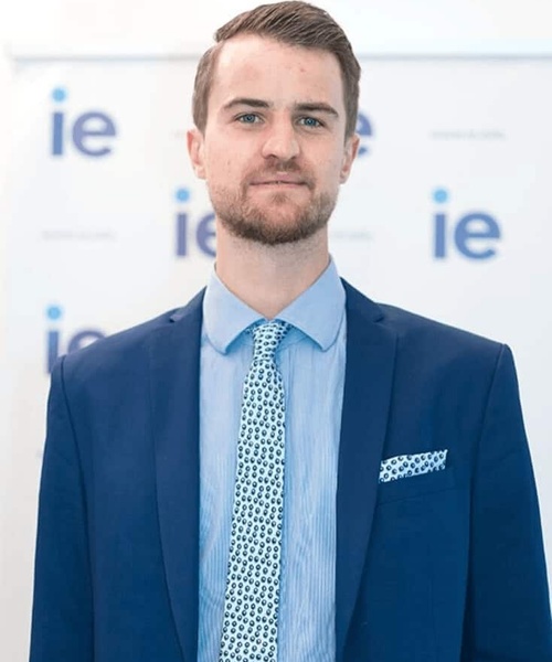 A young man in a business suit with a blue tie and a pocket square standing in front of a background with logos.