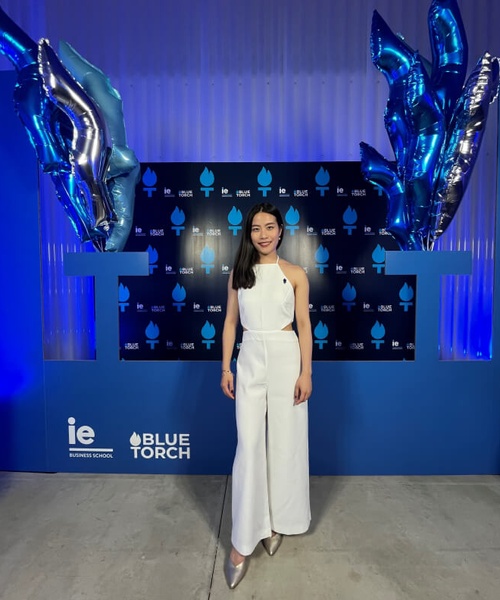 A woman in a white jumpsuit is standing in front of a promotional backdrop with blue balloons and logos.