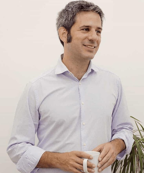 A man in a light blue shirt holding a white mug, smiling and looking to the side.