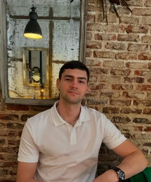 A young man sitting at a table in a rustic cafe setting with a brick wall and hanging lamp reflected in a mirror behind him.