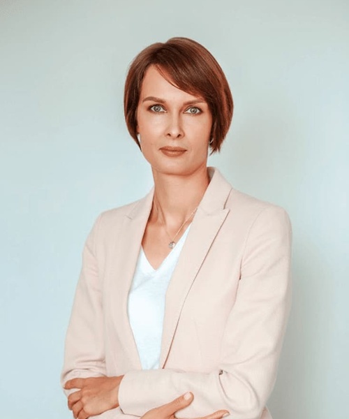 A professional looking woman with short hair stands confidently with her arms crossed, wearing a light pink blazer over a white blouse against a pale blue background.