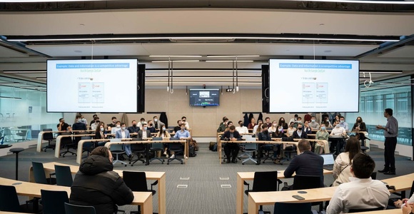 A group of people seated in a modern conference room with presentations displayed on screens.
