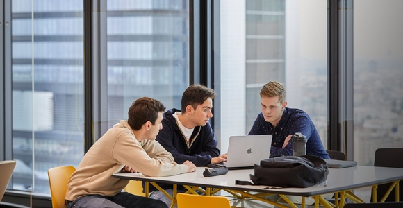 Three young men are collaborating on a laptop at a modern workspace with a city view.