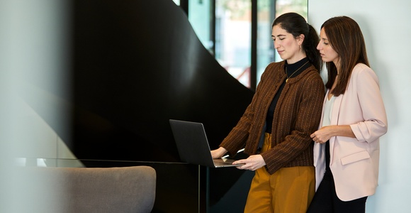 Two women standing and using a laptop together in a modern office setting.