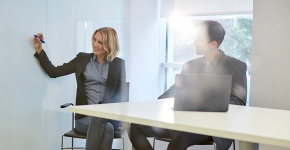 A woman writing on a whiteboard while a man watches, both seated in a modern office setting.
