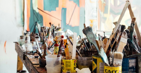 A cluttered artist's table filled with paint brushes, open paint cans, and various painting supplies in front of a colorful abstract painting.