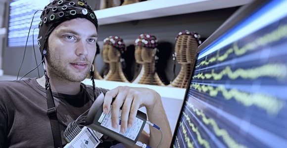 A man wearing an EEG cap observes brainwave readings on a monitor.
