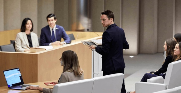 A man is speaking at a podium in a modern conference room with several attendees and a panel of two men and a woman.