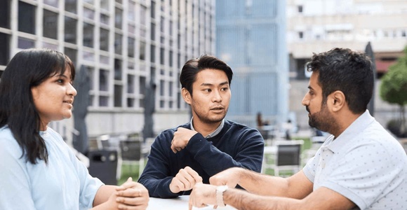 Three people having a conversation at a table outdoors, with modern buildings in the background.