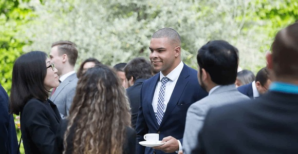 A man in a suit with a tie is smiling while holding a coffee cup, interacting with other people at an outdoor gathering.