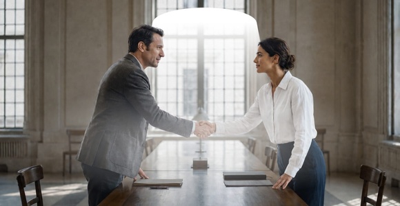 A man and a woman are shaking hands across a table in a bright, spacious room with large windows.