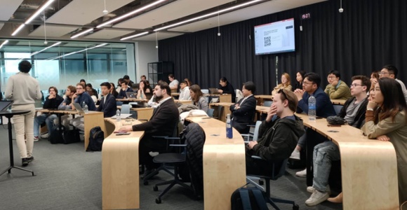 A group of people sitting in a modern classroom listening to a presenter at the front.