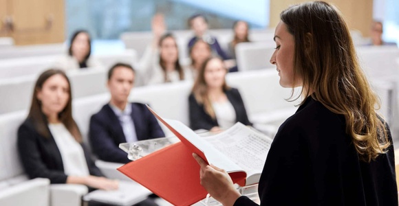 A woman is presenting to an audience in a seminar hall, holding a red folder and addressing attendees who are seated and listening attentively.