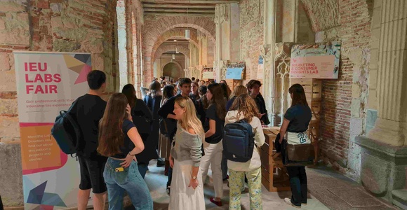 A group of people gathered at an event in a historic building with a stylish banner displaying 'IEU LABS FAIR'.