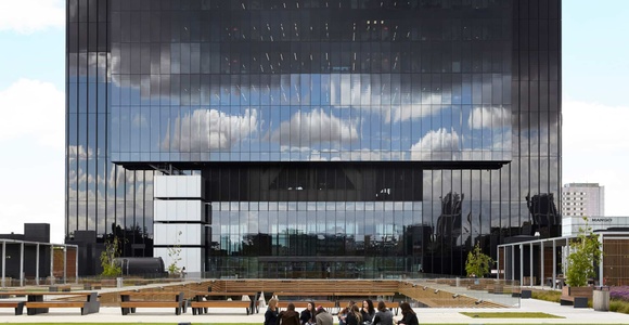 A group of people sitting on the grass in front of a modern glass building with a unique architectural design.