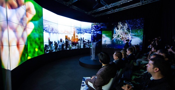People sitting in a dark room watching a panoramic display showing vibrant images of nature and cities.