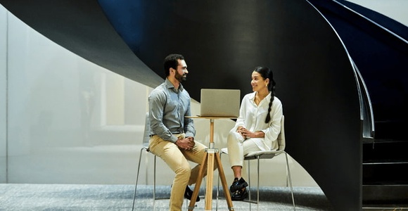 A man and a woman sitting and talking under a large, spiral staircase in a modern building.