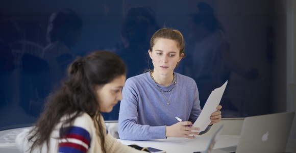 A woman is attentively looking at another woman seated at a table with papers and a laptop.