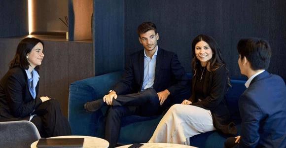 Four business professionals are sitting and engaging in a conversation in a modern office lounge.