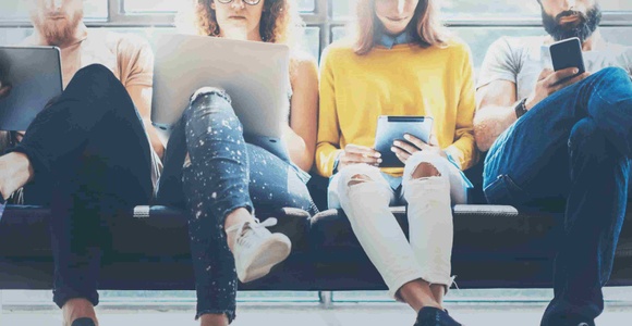 Five young adults sitting on a bench, each engaged with different digital devices.
