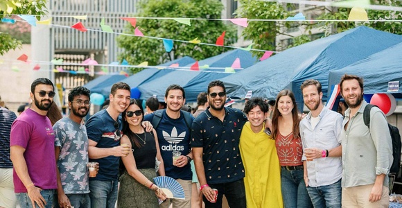 A diverse group of people smiling at an outdoor event with tents and hanging flags.