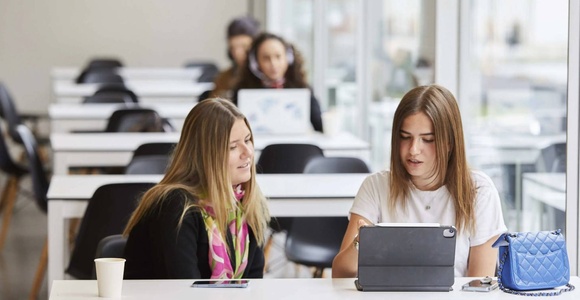 Two women collaborating on a work project with laptops at a white desk in an office environment.