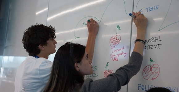 Two individuals are writing on a whiteboard filled with colorful notes and drawings.