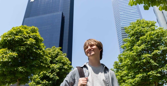 A young man stands smiling in front of modern skyscrapers on a sunny day.