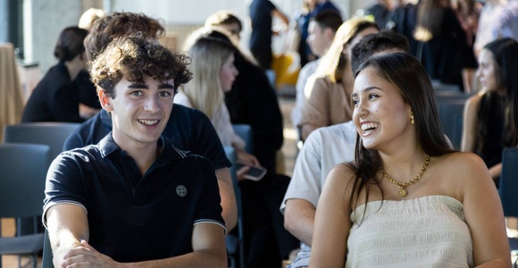 A young man and woman smiling and talking, sitting in a room filled with people.