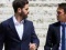 Two men in business suits walking and talking against a white brick wall.