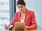 A young woman in a pink blazer is sitting at a table using a tablet while taking notes.