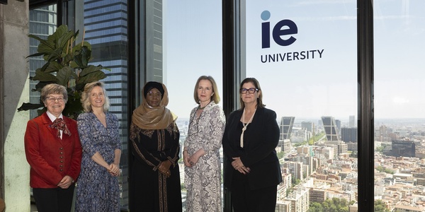 Five women standing in a modern office setting with large windows overlooking a cityscape, next to a sign that reads 'IE University'.