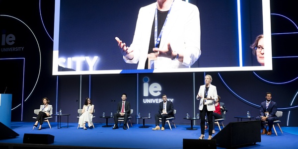 A professional conference event with speakers presenting to an audience, featuring a large screen displaying a close-up of a speaker.