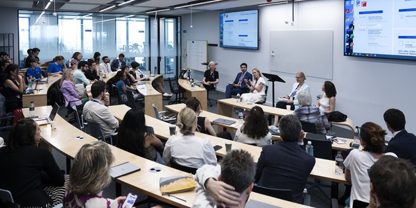 A group of people attending a presentation in a modern conference room with speakers at the front.