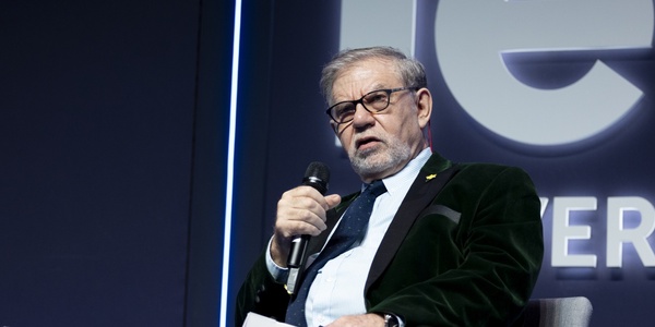 An elderly man with glasses speaking into a microphone at a conference, with a logo visible in the background.