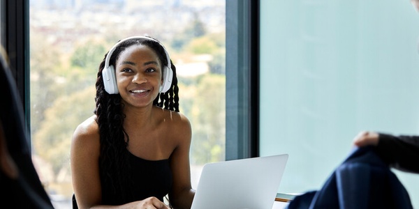 A woman with headphones smiles while working on a laptop in a bright office.