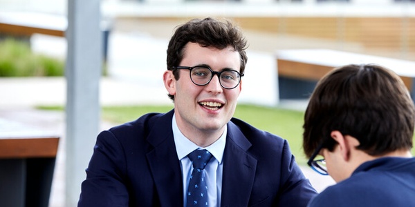 A young man in a suit smiling while sitting at a table, interacting with another person.