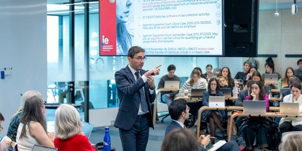 A man gestures while speaking in front of an audience during a presentation.