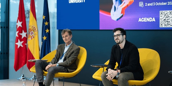 A conference setting with two speakers seated in colorful chairs, discussing in front of a display with flags and event information.