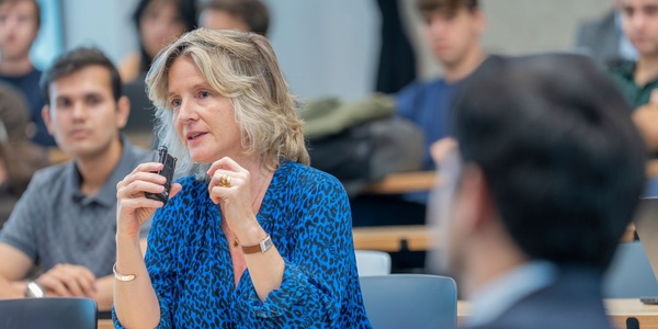 A woman with curly hair is speaking in front of an audience in a classroom setting.
