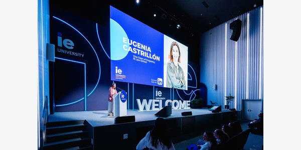 A speaker presenting at an event with a large screen displaying their name and university logo.